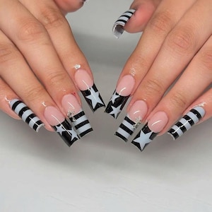 Op de afbeelding: Lange vierkante kunstnagels met een French manicure-stijl op een nude basis. De uiteinden zijn versierd met afwisselende zwarte en witte horizontale strepen en witte sterren. Kleine zilveren kruisbedels zijn aan de nagels bevestigd.