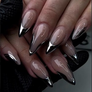Puede incluir: Una manicura con uñas artificiales largas en forma de almendra, con base nude y puntas plateadas cromadas. Las uñas brillantes reflejan la luz, creando un contraste llamativo. La manicura se muestra en una mano con un guante negro.