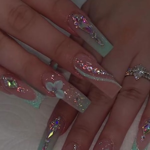 May include: Long, square-shaped acrylic nails with a clear base, adorned with iridescent rhinestones, glitter, and light blue accents. Some nails feature 3D flower embellishments. A diamond ring is also visible.