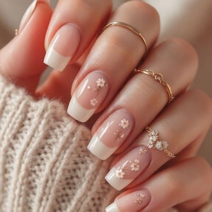 May include: Close-up of a hand with a manicure featuring a French tip design. The nails are adorned with small white flower decals. Gold rings are worn on the fingers, and the hand is resting on a beige knitted jumper.