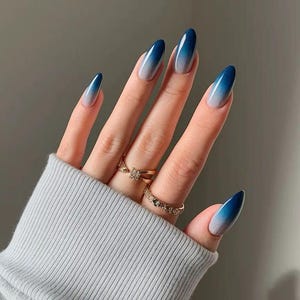Puede incluir: Una mano con uñas largas y ovaladas con un diseño degradado de azul a blanco. Las uñas son brillantes y están bien cuidadas. Anillos dorados adornan los dedos, y la mano está parcialmente cubierta por una camisa de manga larga gris claro.