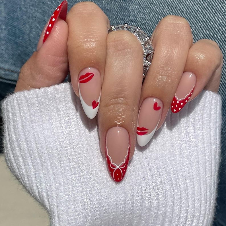 May include: A hand with almond-shaped nails featuring a Valentine's Day theme. The nails are painted with a nude base and white tips, adorned with red lips, hearts, and polka dots. One nail is entirely red with white polka dots.