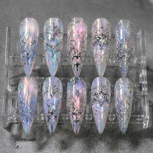 May include: A set of ten stiletto-shaped press-on nails featuring an iridescent, pearlescent finish with raised, metallic silver 3D liquid-style filigree patterns. The nails are arranged in two rows on a clear plastic display stand.