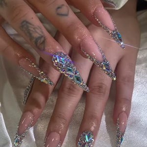 May include: Long, pointed acrylic nails with a nude base and sparkling silver and iridescent crystal embellishments. The nails are adorned with various sizes and shapes of crystals, creating a glamorous and eye-catching design.