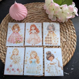 Op de afbeelding: Set van zes rechthoekige kunstprints met prinsessenthema. Elke print toont een andere illustratie van een prinses in een jurk, met details zoals bloemen, een lantaarn en een eenhoorn. Een roze kaars en een bloemstuk zijn ook zichtbaar.