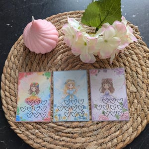 Op de afbeelding: Drie kaarten met feeënthema's met hartvormige contouren, een roze kaars en een tros witte en roze bloemen op een geweven mat. De kaarten bevatten illustraties van feeën in kleurrijke jurken en bloemaccenten.