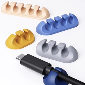 Könnte beinhalten: Eine Auswahl an Kabelhaltern in Beige, Blau, Gelb und Grau. Jeder Halter hat mehrere Schlitze zur Aufnahme von Kabeln. Ein schwarzes USB-C-Kabel wird in einen blauen Halter eingesetzt, um die Verwendung zu demonstrieren.