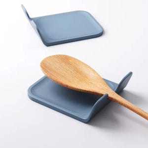 Può includere: Sono mostrati due poggiacucchiaio blu-grigi, uno vuoto e uno con un cucchiaio di legno. I poggiacucchiaio hanno una forma rettangolare con un fronte curvo e un retro rialzato per tenere le posate. Il cucchiaio di legno è marrone chiaro.