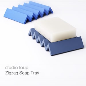 Könnte beinhalten: Zwei blaue Zickzack-Seifenschalen, eine mit einem weißen Seifenstück. Die Schalen haben ein modernes, geometrisches Design. Der Text "studio loup Zigzag Soap Tray" ist sichtbar. Hergestellt aus einem festen Material.