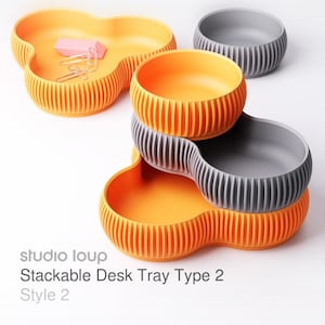 Könnte beinhalten: Orange und graue, stapelbare Schreibtischablagen mit geripptem Design. Eine Ablage hat eine kleeblattartige Form und enthält einen rosa Radiergummi und Büroklammern. Der Text "studio loup Stackable Desk Tray Type 2 Style 2" ist sichtbar.