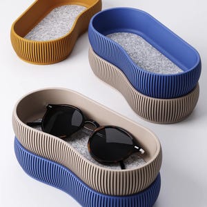 Può includere: Tre vassoi portaoggetti ovali impilati, nei colori giallo senape, blu e beige. Ogni vassoio ha i lati a coste verticali e un interno in feltro grigio. Gli occhiali da sole poggiano nel vassoio beige. I vassoi sono progettati per organizzare piccoli oggetti.