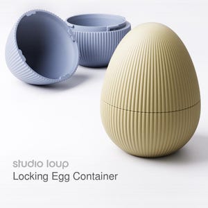 Könnte beinhalten: Ein hellgelber, eiförmiger Verschlussbehälter mit vertikalen Rippendetails. Im Hintergrund sind zwei blaue Behälter zu sehen. Der Text "studio loup Locking Egg Container" ist sichtbar.