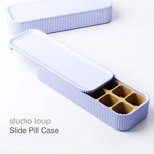 Könnte beinhalten: Eine hellviolette Schiebe-Pillenbox mit gerippter Außenseite und kontrastierendem goldfarbenem Innenraum. Die Box ist rechteckig mit einem Schiebedeckel. Der Text "studio loup Slide Pill Case" ist auf der Seite aufgedruckt.