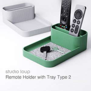 Könnte beinhalten: Ein grüner und ein grauer Fernbedienungshalter mit Ablage. Der grüne Halter hat zwei Fernbedienungen und Schlüssel in der Ablage. Der graue Halter ist leer. Der Text "studio loup Remote Holder with Tray Type 2" ist im Bild.