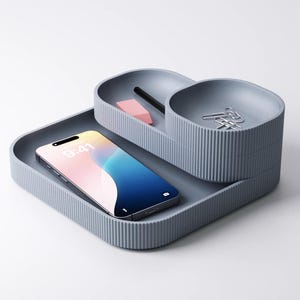 Könnte beinhalten: Ein grauer Schreibtisch-Organizer mit gerippter Textur. Er hat drei Fächer: ein rechteckiges Tablett mit einem Smartphone mit der Anzeige "9:41", einen kleineren ovalen Bereich mit einem Bleistift und einem Radiergummi sowie einen runden Bereich mit Büroklammern. Der Organizer befindet sich auf weißem Hintergrund.