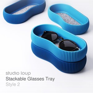 Könnte beinhalten: Drei blaue, stapelbare Brillenablagen mit strukturierter Außenseite und grauem Innenraum. Eine Ablage enthält eine Sonnenbrille. Der Text "studio loup Stackable Glasses Tray Style 2" ist sichtbar.