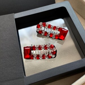 May include: Two red and silver barrettes with a rectangular shape, adorned with sparkling red and clear crystals. The barrettes are displayed in a black box with a reflective surface, suggesting a jewelry or accessory item.