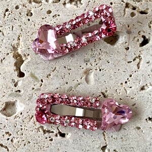 May include: Two pink hair clips with heart-shaped accents. Each clip is adorned with small, sparkling rhinestones and a larger heart-shaped gem. The clips are set against a textured, light-colored surface.