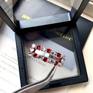 May include: A rectangular hair clip with a red base, adorned with clear and red crystals. The clip is displayed in a black box with a clear lid, held by tweezers. The text "PAWSTUDIO" is visible.