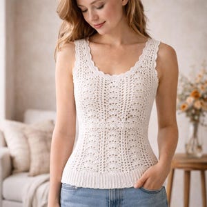 May include: A cream-coloured crochet vest top with a scalloped neckline and straps. The top has a detailed openwork pattern and a ribbed hem. The sleeveless garment appears handmade, showcasing a delicate and textured design. The top is likely made of cotton.