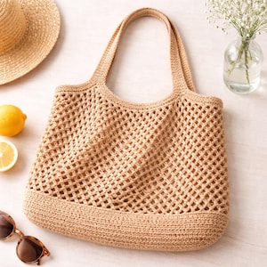 May include: A beige crochet tote bag with two handles. The bag has a net-like design and a solid base. A straw hat, sunglasses, and a lemon are next to the bag. A small glass vase with white flowers is in the background.