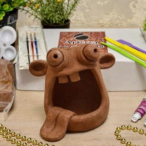 May include: A whimsical brown clay sculpture with a wide open mouth and protruding tongue is the focal point. The image also includes a craft kit, paint tubes, sculpting tools, and decorative beads, suggesting a creative project.