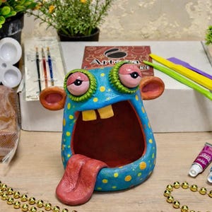 May include: A whimsical, handmade ceramic monster sculpture with a wide open mouth and a long red tongue. The blue body is decorated with yellow dots. The sculpture is part of a clay art kit, with tools, paints, and clay visible in the background.