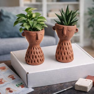 May include: Two terracotta-colored face planters, each holding a different houseplant. The planters have closed eyes and smiling mouths. The base of each planter has a decorative cut-out pattern. A white box, clay, and instructions are in the foreground.