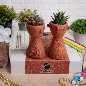 May include: Two terracotta-colored face planters, each holding a succulent plant, sit on a box with the Arteza logo. The box contains art supplies, including paint tubes, paintbrushes, and a palette. Decorative gold beads and small plants surround the box.