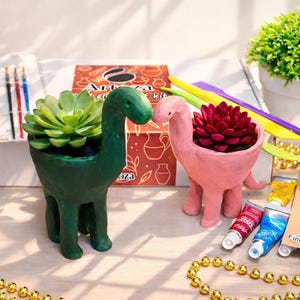 May include: A green and a pink dinosaur-shaped planter, each holding a succulent plant. The green planter has a light green succulent, and the pink planter has a dark red succulent. Craft supplies and a string of gold beads are also visible.