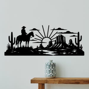 May include: A black metal wall art piece featuring a silhouette of a cowboy on horseback, cacti, and a rising sun over a desert landscape with mesas. The design includes clouds and a flat base, creating a wide, horizontal decorative scene.
