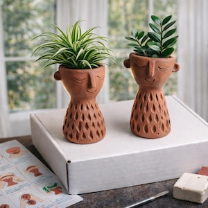 May include: Two terracotta-colored face planters, each holding a different green plant, sit on a white box. The planters have closed eyes and smiling mouths. A craft kit with clay and tools is nearby.