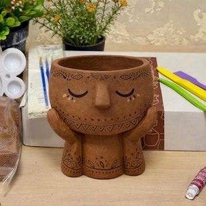 May include: A brown ceramic planter with a face design, featuring closed eyes and hands cradling the sides. The planter has decorative patterns and is surrounded by art supplies, including paint tubes and brushes. The overall theme is arts and crafts.
