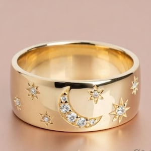 May include: A wide, gold-colored ring featuring a crescent moon and star design. The moon is adorned with small, clear gemstones, and stars are scattered around the band. The ring has a polished finish.