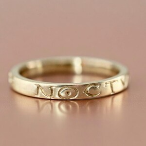 May include: A gold ring with the word "NOCTY" engraved around the band. The ring has a smooth, polished finish and is set against a soft, rose-coloured background. The ring is a piece of jewellery.