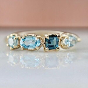 May include: A gold ring featuring four blue gemstones. The gemstones are various shapes, including round, oval, and square. The ring is set against a soft pink background.