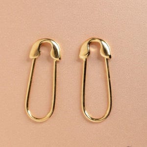 Op de afbeelding: Een paar goudkleurige veiligheidsspeld oorbellen. De oorbellen worden gepresenteerd op een lichtroze achtergrond. De oorbellen zijn gemaakt van een glanzend, metalen materiaal en hebben een klassiek veiligheidsspeldontwerp.