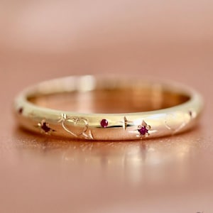 May include: A gold-colored ring with engraved heart, star, and arrow designs, interspersed with small red gemstones. The ring is set against a soft, pink background, highlighting its delicate details and craftsmanship.