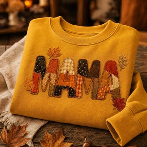 Peut inclure: Un sweat-shirt à col rond jaune moutarde avec le mot MAMA appliqué dans un style patchwork. Les lettres sont confectionnées à partir de divers tissus dans des tons orange, marron et crème avec des coutures visibles. Des feuilles d'automne brodées ornent le motif.