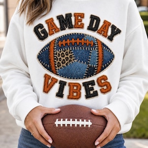 Könnte beinhalten: Ein weißes Sweatshirt mit Rundhalsausschnitt und dem Schriftzug GAME DAY VIBES in orangefarbenen und schwarzen Buchstaben im Varsity-Stil. Mittig befindet sich eine Grafik eines Footballs mit blauer Denim-Textur, Leopardenmuster und orangefarbenen Nähten.