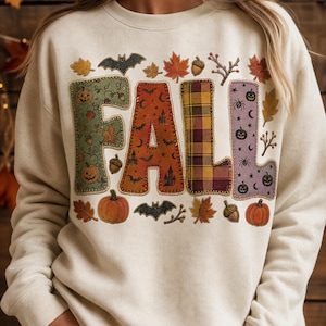Op de afbeelding: Een crèmekleurige sweater met ronde hals en het woord FALL in grote blokletters. Elke letter heeft een uniek patroon: groen met pompoenen, oranje met vleermuizen, rood-geel geruit en paars met spinnen en lachende pompoenen.