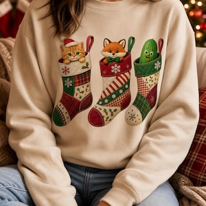 Op de afbeelding: Een beige sweater met ronde hals en een grafische print van drie kerstsokken. De eerste sok bevat een oranje cyperse kitten met een kerstmuts, de tweede een vos met een groene vlinderdas en de derde een lachende groene augurk.
