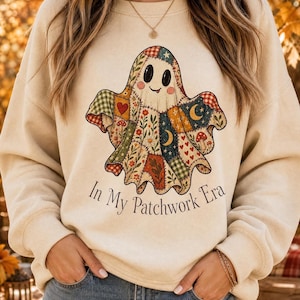 Puede incluir: Una sudadera beige de cuello redondo con un gráfico central de un fantasma hecho de varios patrones de patchwork coloridos que incluyen corazones, setas, estrellas y lunas. El texto "In My Patchwork Era" está impreso en negro debajo.