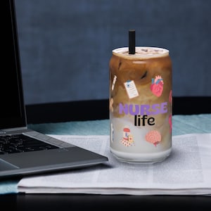 Könnte beinhalten: Ein durchsichtiger, dosenförmiger Becher, gefüllt mit einem geschichteten Eiskaffee und einem schwarzen Strohhalm. Der Becher ist mit medizinischen Aufklebern und den Worten "NURSE life" in Lila verziert.