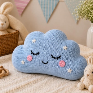 May include: A light blue cloud-shaped pillow with a crocheted texture. The pillow features embroidered closed eyes with long lashes, a small curved smile, two pink circular cheeks, and several small white stars scattered across the surface.