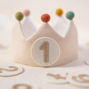 Könnte beinhalten: Eine beigefarbene Stoffkrone mit einem runden Aufnäher, der eine goldene Glitzerzahl 1 zeigt. Die Krone ist mit fünf kleinen Filz-Pompons in Olivgrün, Hellrosa, Orange, Gelb und Blaugrün verziert. Das Innere ist mit weißem Stoff ausgekleidet.