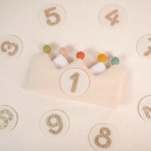 Peut inclure: Une couronne en feutrine beige avec un écusson circulaire central affichant le chiffre 1 en paillettes dorées. La couronne est surmontée de cinq petits pompons colorés. Des disques en feutrine avec les chiffres 2 à 9 dorés entourent la couronne.