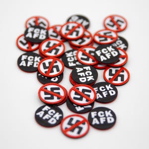 Könnte beinhalten: Eine Ansammlung kleiner, runder schwarzer Knöpfe mit roten Rändern. Jeder Knopf zeigt ein weißes Hakenkreuz-Symbol, das mit einer roten Linie durchgestrichen ist, und den weißen Text "FCK AFD". Die Knöpfe liegen auf einer weißen Oberfläche.