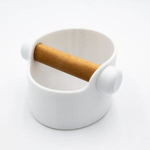 Könnte beinhalten: Eine weiße Kaffeemehl-Abschlagbox mit einer Holzstange. Die Abschlagbox hat eine zylindrische Form mit einer gebogenen Öffnung und zwei weißen, abgerundeten Griffen. Die Holzstange ist 15 cm lang.