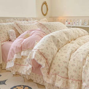Puede incluir: Una cama con un edredón y fundas de almohada con estampado floral en crema y rosa. La ropa de cama tiene bordes con volantes y una falda de cama rosa. Un pequeño oso de peluche y una taza están en la cama.